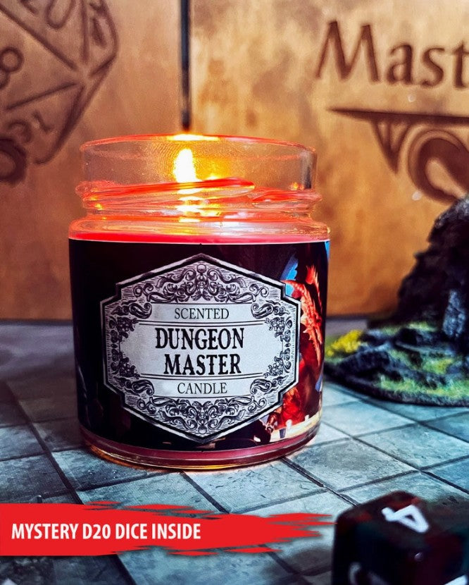 A lit scented candle in a clear glass jar labeled 'Scented Dungeon Master Candle' sits on a stone-tiled surface with a wooden background and gaming-themed props, including a red banner reading 'Mystery D20 Dice Inside'.