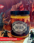 A 'Scented Tavern Candle' in a clear glass jar with a black lid, featuring a label with fantasy artwork. The candle is displayed on a tabletop with miniature figurines, with a red banner overlay at the bottom reading 'INCLUSIVE SUPRISE D20 INSIDE'.