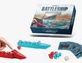 Farnell Battleship search and destroy game set with its retail box, a red player ship, a blue player ship, red and blue dice, and cards on a white background.